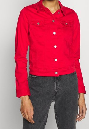 Kvinde iført en knap-op lyserød jakke med brystlommer og sorte jeans med høj talje, stående mod en ensfarvet baggrund.