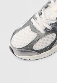 Γκρι και λευκό αθλητικό παπούτσι με επάνω μέρος από δίχτυ και συνθετικό υλικό, διαθέτοντας μαξιλαρωμένη γλώσσα, στρογγυλεμένη μύτη και υφή στη σόλα.