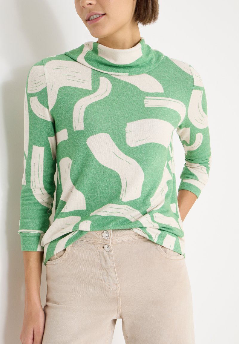 Haut vert à manches longues avec des motifs abstraits en coups de pinceau blancs; texture douce avec un col haut, associé à un pantalon beige.