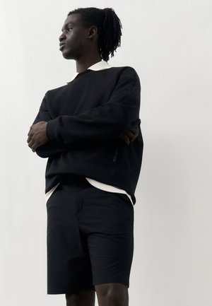 Mladý muž s dredmi, ktorý má na sebe čiernu mikinu cez bielu košeľu s golierom a čierne šortky, stojí so skríženými rukami na jednoduchom pozadí.