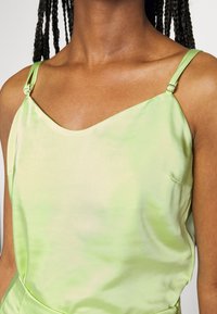 Camisola de cetim verde claro com alças ajustáveis, apresentando uma textura suave e um padrão de tie-dye subtil, vista da área do ombro.