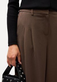 Brune skræddersyede bukser med folder, parret med en struktureret sort håndtaske prydet med sølvnitter, og en ribbet sort sweater.
