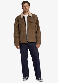 Chaqueta de pana marrón con un cuello de felpa beige, dos bolsillos en el pecho y cierre de cremallera en la parte delantera, combinada con jeans oscuros y zapatillas.