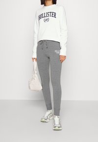 Vit sweatshirt med "Hollister"-logotyp, grå tajta sweatpants med sidologotyp, bär en beige väska och har på sig silverfärgade träningsskor.