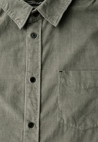 Camisa de pana verde con un cuello puntiagudo, cinco botones negros y un bolsillo en el lado izquierdo del pecho. La textura es acanalada con un acabado suave.