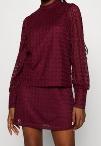 Set a due pezzi in pizzo borgogna composto da una maglia a maniche lunghe con colletto alto e una minigonna aderente, con motivo a rombi testurizzati su tutta la superficie.