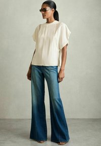 Cremefarbene Bluse mit Spitze und kurzen Ärmeln, kombiniert mit blauen, weit geschnittenen Jeans mit sichtbaren Nähten. Glatte Textur aus leichtem Stoff.