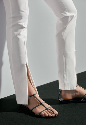 Parties basses des jambes portant un pantalon blanc avec des fentes latérales et des sandales plates noires à brides sur un sol sombre contre un mur clair.