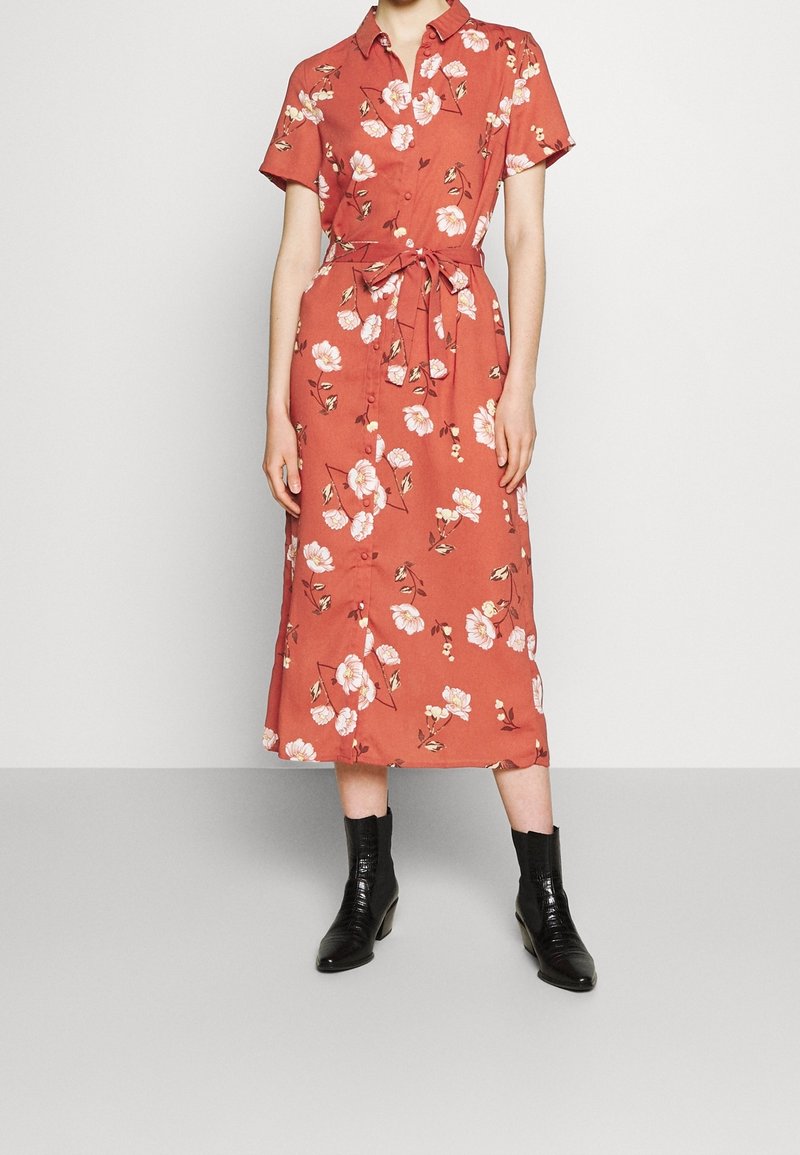 Femme portant une robe midi fleurie à manches courtes couleur rouille avec des boutons et une ceinture nouée à la taille, associée à des bottines noires.
