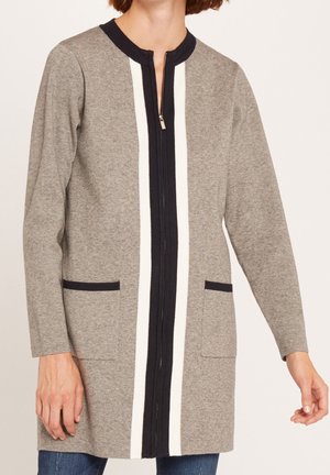 Femme portant un cardigan gris à manches longues avec des rayures verticales noires et blanches et des poches avant, associé à un jean bleu.
