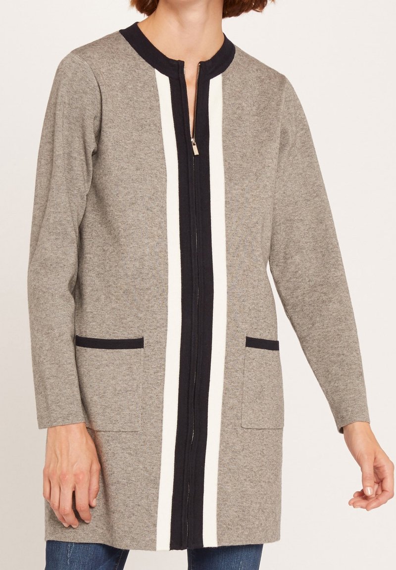 Femme portant un cardigan gris à manches longues avec des rayures verticales noires et blanches et des poches avant, associé à un jean bleu.