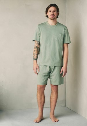 Man standing barefoot in a green short-sleeve shirt and matching shorts with a tattoo sleeve on his left arm against a plain wall.