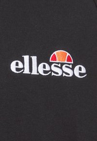 Logo Ellesse brodé sur tissu noir, avec texte blanc en minuscules et un motif en demi-cercle rouge orangé au-dessus des lettres.