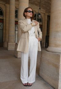 Beige linen blazer over a white blouse, paired with high-waisted white trousers and black flip-flops. Gold jewelry accents the outfit.