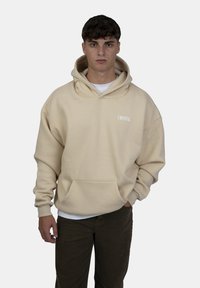 Felpa oversize beige con tasca frontale, cappuccio con cordino e testo bianco "CRYSTAL" sul lato sinistro del petto. Indossata sopra una maglietta bianca.