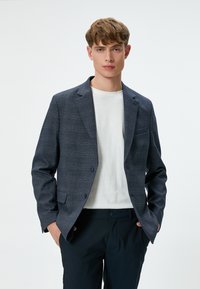 Marinblå rutig kavaj i strukturerad tyg, med snedskuren krage, två knappar och framfickor, bärs över en vit T-shirt med rund hals.