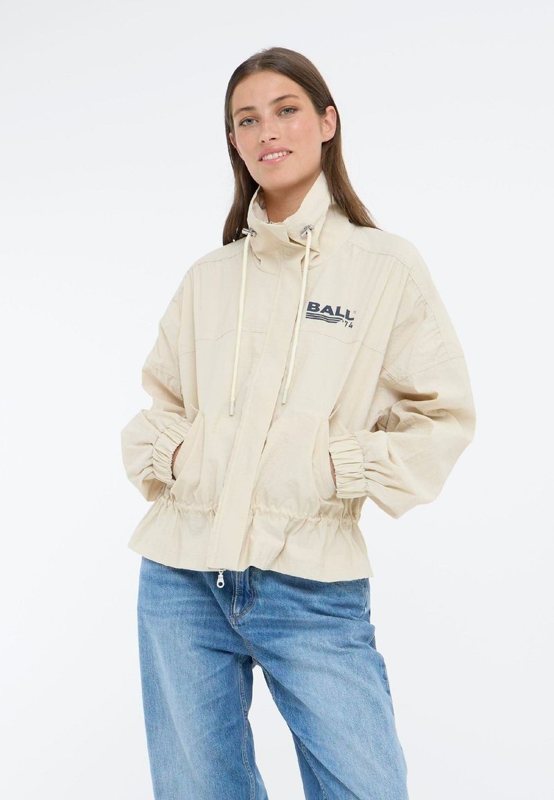 Veste légère beige avec un col haut, fermeture éclair, poignets élastiques et taille à cordon de serrage. Présente le logo "BALL" imprimé en bleu fonc�é.