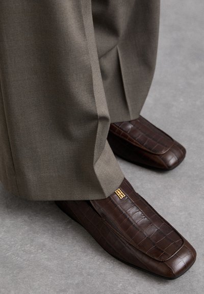 Brune læder loafers med krokodillemønster, prydet med en gylden logoaccent, parret med lysegrå bukser. Kvadratisk tådesign.