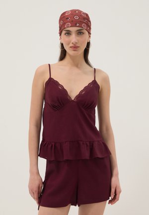 Mujer con camisola burdeos con encaje y pantalones cortos a juego con bandana de color rojizo con estampado y pendientes de aro.