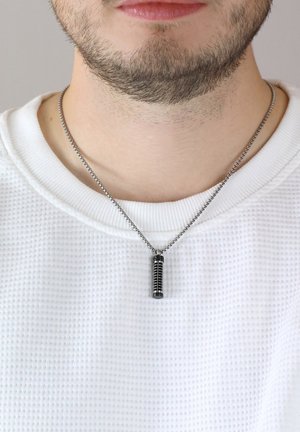 Silberne Kettenhalskette mit einem zylindrischen Anhänger, der schwarze Rillen und eine glatte Oberseite aufweist, vor einem strukturierten weißen Stoff.
