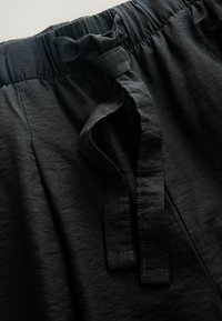 Pantalon en tissu noir avec taille élastique et cordon noué à l'avant, montrant la texture et les détails de couture.