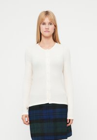 Cardigan alb cu dungi, cu guler rotund și nasturi în față. Mâneci lungi și textură moale. Purtat peste o fustă în carouri închise la culoare.