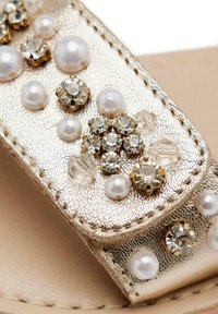Tira de sandalia metálica dorada que presenta piedras de rhinestone transparentes de diversos tamaños y perlas blancas, con un acabado texturizado y detalles de costura.