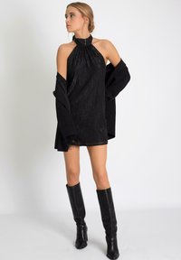 Robe noire à col halter avec une texture plissée et un ourlet à franges, associée à une veste noire surdimensionnée et des bottes à talons noires jusqu'aux genoux.