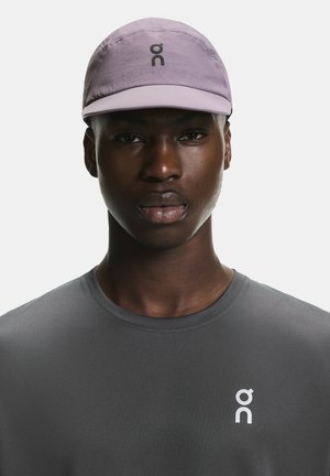 Hellviolette Mütze mit weicher Textur und gebogenem Schirm; mit einem schwarzen Logo. Das Modell trägt ein enganliegendes graues Sportshirt mit rundem Ausschnitt.