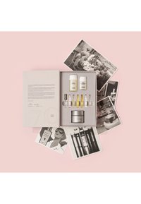 Set de regalo de cuidado de la piel en una caja gris claro con dos botellas blancas, varias ampollas de vidrio y un frasco plateado. Rodeado de fotos en blanco y negro.