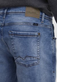 Jeans de mezclilla azul claro con un diseño de bolsillo distintivo, costuras en ángulo y una etiqueta de la marca en la cintura trasera, que muestra un acabado texturizado.