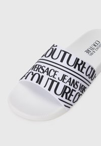 Baltas šlepetes, pagamintas iš tekstūruoto gumos, su plačia juosta, ant kurios yra juodi tekstiniai raštai, įskaitant "Versace Jeans Couture."
