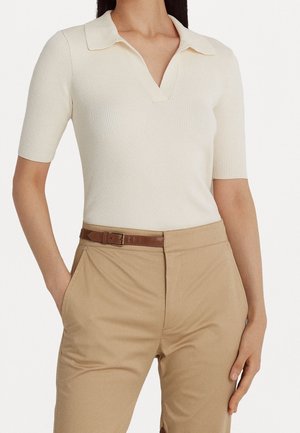 Femme portant un polo en maille côtelée crème à manches courtes, rentré dans un pantalon beige avec une ceinture en cuir marron, mains détendues le long du corps.