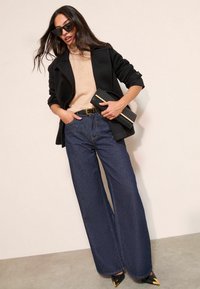 Leveälahkeiset tummansiniset farkut yhdistettynä beigeen neuleeseen ja mustaan takkiin. Asustettu mustalla clutch-laukulla ja mustilla teräväkärkisillä koroilla.