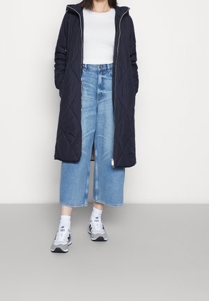 Personne portant un long manteau noir matelassé sur un haut blanc côtelé, un jean bleu large et court, des chaussettes blanches et des baskets grises.