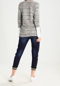 Grauer gemusterter Pullover mit langen weißen Ärmeln, kombiniert mit dunkelblauen, schmalen Jeans mit gestreiften gelben Bündchen. Weiße Sneaker.