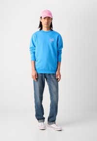 Man in een felblauwe sweatshirt met roze tekst, lichtroze pet, blauwe jeans met cuffs en witte sneakers staand tegen een effen witte achtergrond.