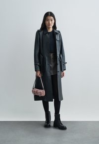 Trench coat in pelle nera con vita stretta, abbinato a una maglia nera aderente, gonna marrone, leggings neri e stivali con lacci. Accessoriato con una piccola borsa rosa.