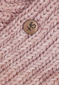 Handstickad mjuk rosa tyg med ett strukturerat mönster, med en cirkulär bronsknapp där bokstäverna "YS" är präglade.