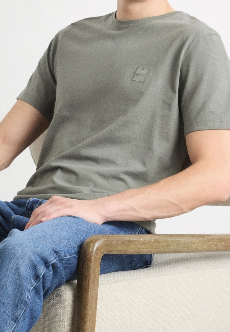 Homme portant un T-shirt Boss vert olive et un jean bleu, assis sur une chaise beige avec accoudoir en bois, sur un fond uni.
