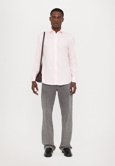 Camicia rosa chiaro con bottoni, maniche lunghe; indossata con jeans a gamba larga grigi e scarpe nere. Piccola borsa a tracolla scura. Sfondo semplice.