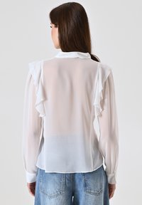 Blusa bianca trasparente con dettagli arricciati sulle spalle, maniche lunghe e colletto classico. Tessuto leggero con una texture liscia.