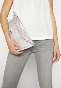 Bolso de mano con cuentas plateadas y una correa de tela anudada, que presenta un brillo metálico y una textura suave, sostenido en una mano contra unos jeans grises.