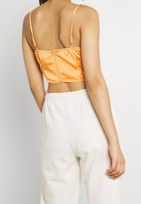 Osoba stojící zády má na sobě oranžový crop top s tenkými ramínky a bílé tepláky, čímž vystihuje minimalistický styl.