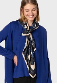 Niebieski sweter robiony na drutach z długimi rękawami, z trójkątnym czarno-kremowym szalem w kwiatowy wzór, noszonym luźno wokół szyi.