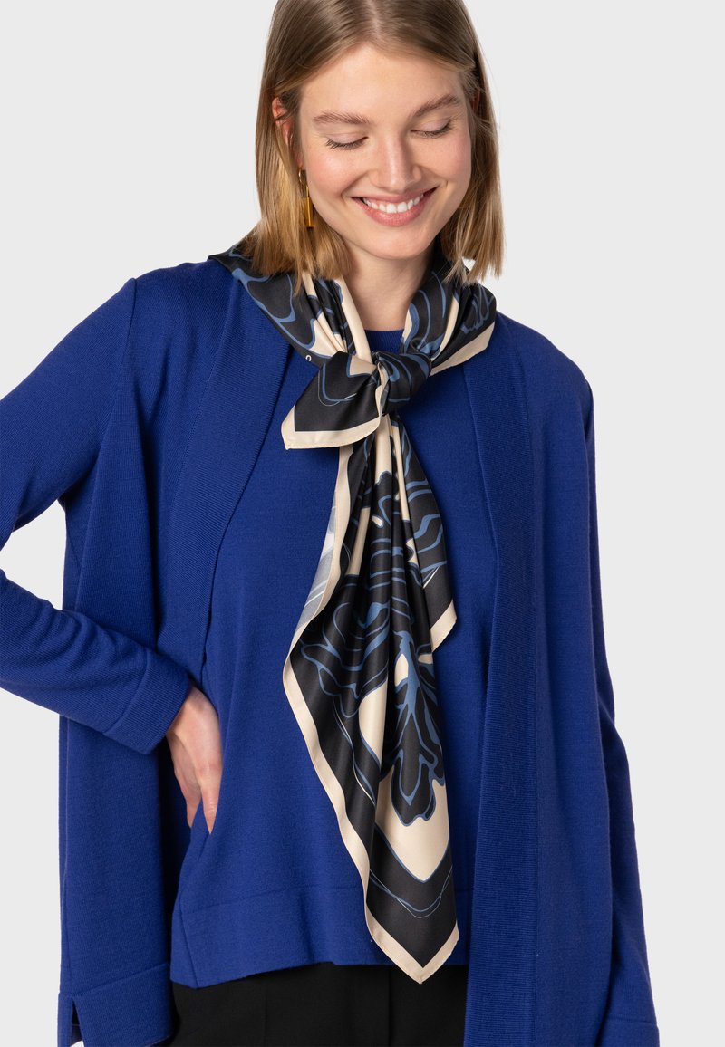 Niebieski sweter robiony na drutach z długimi rękawami, z trójkątnym czarno-kremowym szalem w kwiatowy wzór, noszonym luźno wokół szyi.