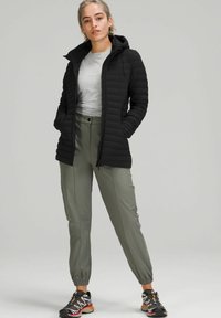 Zwarte pufferjack met capuchon, gecombineerd met een lichtgrijze shirt en olijfgroene tapered joggingsbroek. Zwarte sportschoenen met kleurrijke accenten.