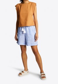 Brun bomulds kortærmet top sat sammen med blå og hvide stribede shorts med hvide frynsedetaljer. Beige sandaler fuldender outfittet.