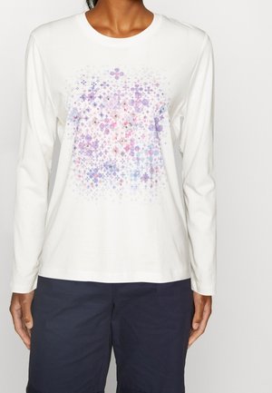 Person iført en hvid langærmet skjorte med et pastelfarvet mønster af lyserøde, lilla og blå plusser og cirkler på forsiden.