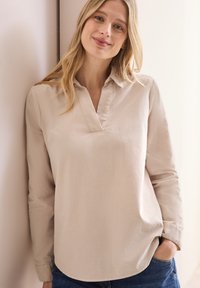 Blusa beige de mangas largas con una tapeta de media botonera y cuello tipo camisa, que presenta un corte relajado y una textura suave sobre un fondo claro.